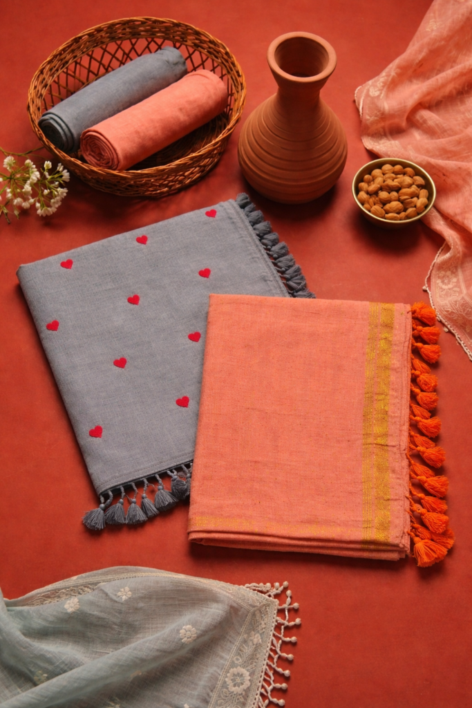 Grey linen saree with red heart motifs and peach saree with gold border styled on terracotta background