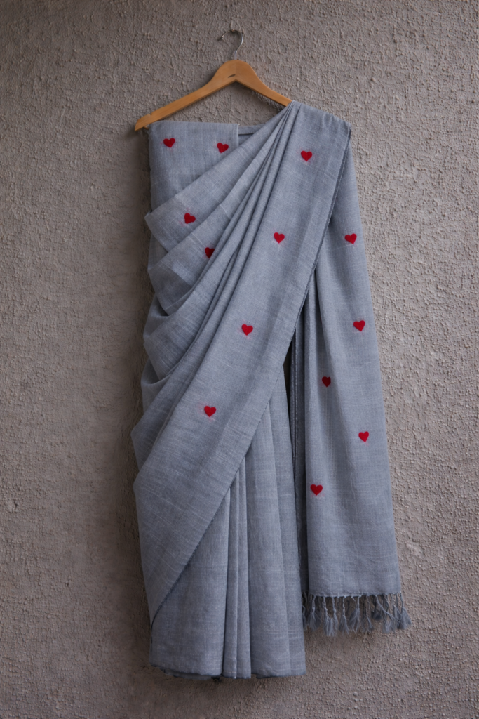 A soft grey linen saree draped on a wooden hanger against a textured wall, featuring small red heart embroidery scattered across the fabric and a subtle striped border with tassels.